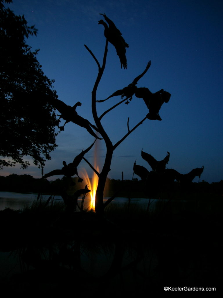 Just after sunset at the Chicago Botanic Garden we see the silhouette of a fowl sculpture depicting what appears to be ducks in midflight entering and leaving the reflecting pond in the background. The pond has a fountain streaming high in to the air with a single light at the bottom to help backlight the metal sculpture against the twilight sky.