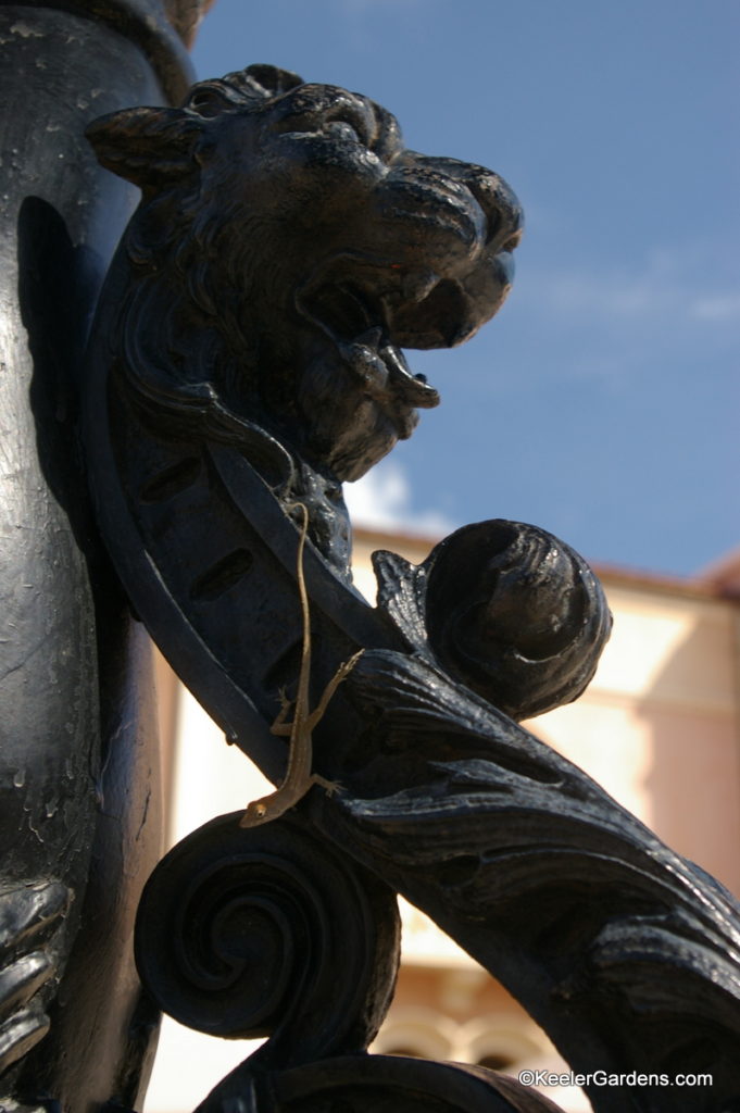 Metal lion heads adorn this decorative base to a light pole. The black metal iron is a popular place for these little salamanders to sun themselves. This one freezes after trying to make a break for it hoping no one notices a grey lizard on a black metal lion.