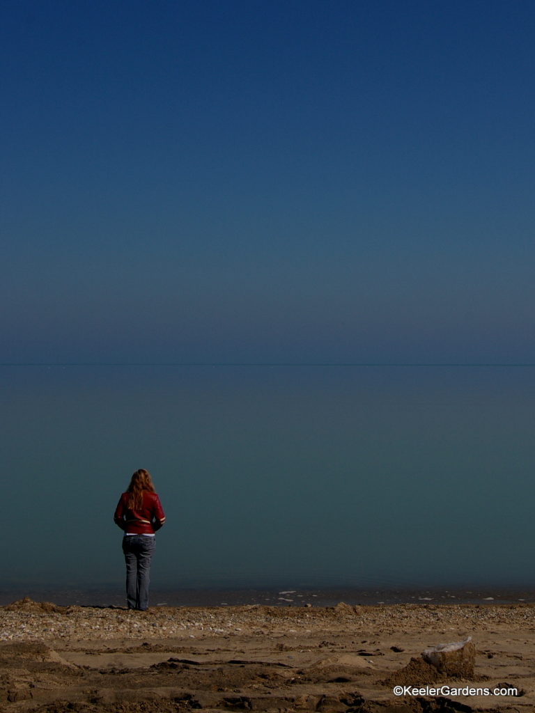 A lone visitor to a somewhat rocky beach in Lake Bluff, IL stands near the water's edge on peers out to an apparently infinite expanse that is Lake Michigan that at some point meets the sky in a thin blue line.