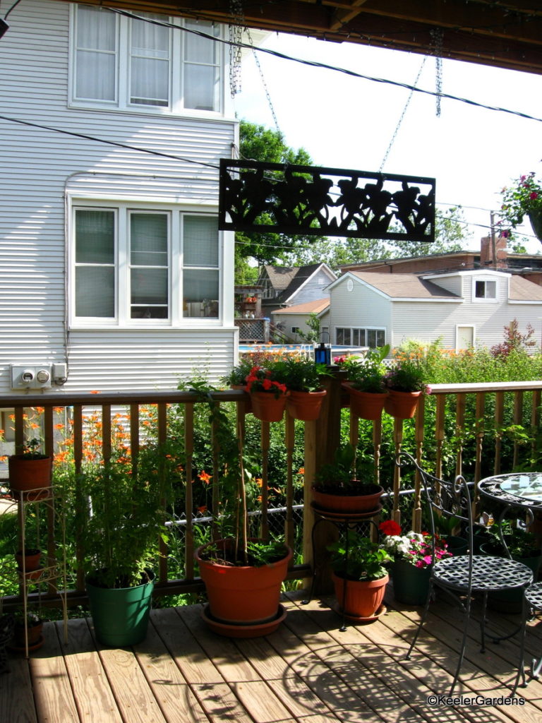 The view on the deck at Keeler Gardens shows what appears to be a metal art piece hung from two chains. The black iron serves as a great contrast against the neighboring white house. Around the deck are various plantings of petunias, impatiens, geraniums, lily of the valley, and marigolds.