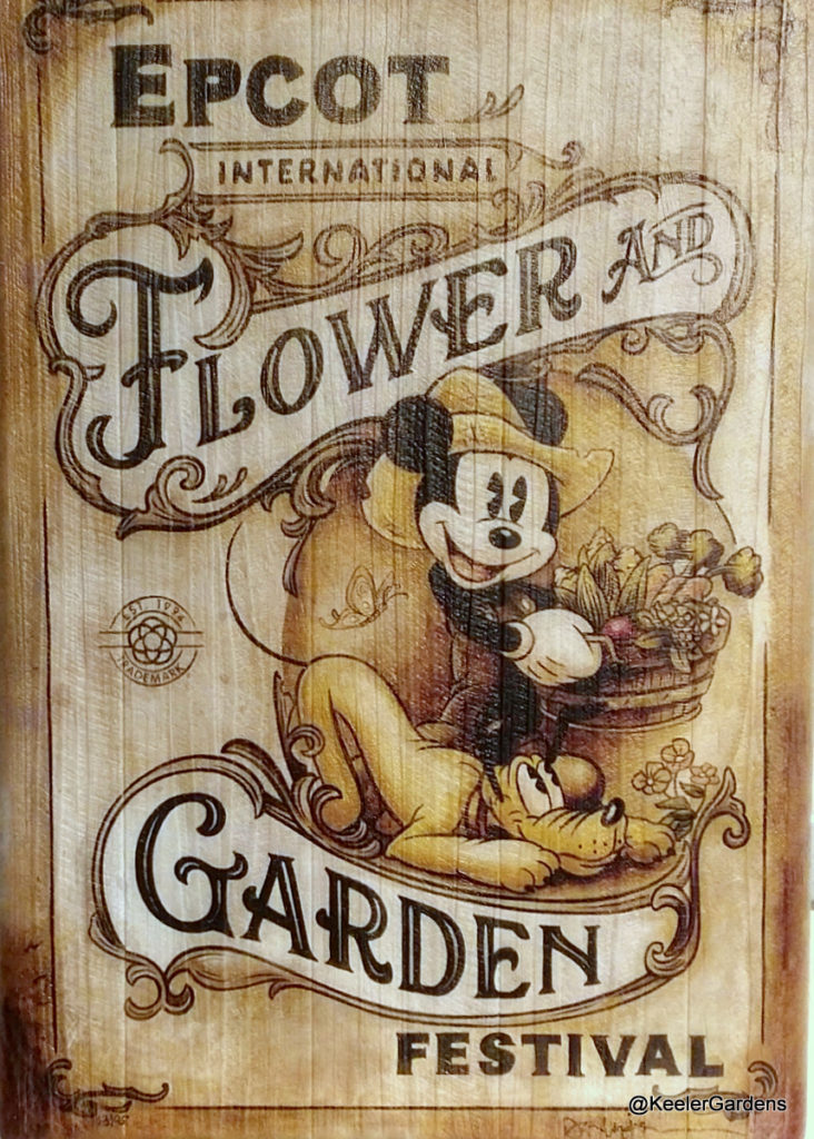 A white washed wooden sign for the Epcot International Flower and Garden Festival showing Mickey Mouse in overalls and a farm hat carrying what appears to be a buschell of corn tulips and asperagas while pluto eagerly looks on under foot.